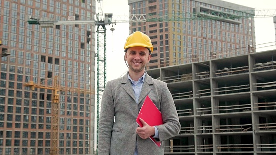 Joyous construction auditor with clipboard under arm standing among unfinished multi-story houses. Dolly shot