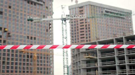 Closeup of red-and-white barrier tape marking off construction zone with tower cranes and unfinished multi-story houses. Slow motion