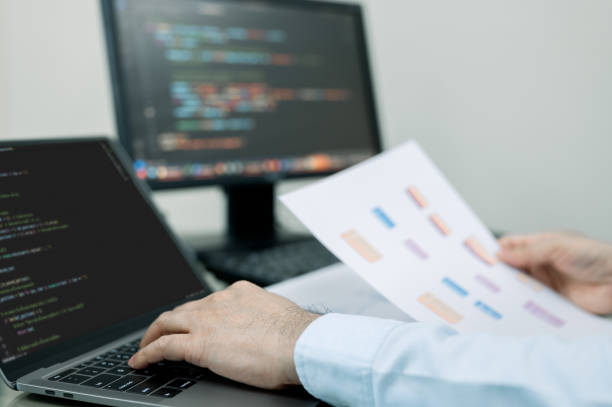 istock programmer is typing on a laptop computer while holding a piece of paper. Laptop is open to a screen displaying code, and the man is focused on his work. Concept of concentration and productivity 2138295859