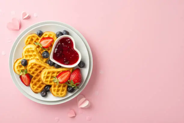 Mother's Day breakfast scene: top view showcasing a plate of heart-shaped waffles, ripe strawberries, blueberries, jam, and paper hearts on a pastel pink backdrop with space for wording