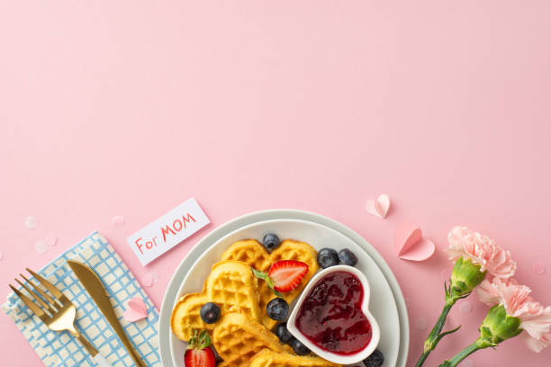 istock Mother's Day breakfast idea: top view heart-shaped waffles, sliced strawberries, blueberries, jam, napkin, utensils, pink carnations, note 