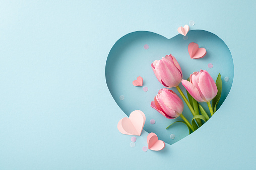 Mother's Day finesse concept. Top view shot of blooming tulips, decorative hearts, modern confetti, showcased within heart-shaped cutout on chic blue background, leaving space for customized promotion