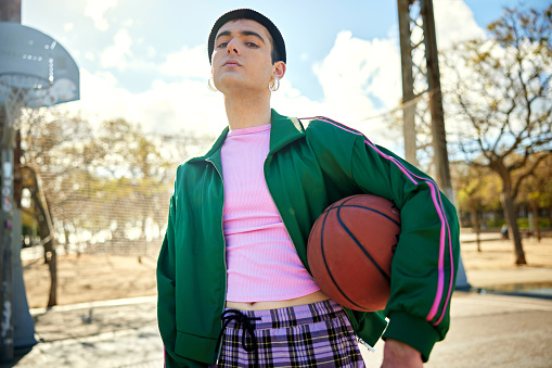 Waist-up view of early 20s man in colorful sports clothing, ball under arm, head tilted up and looking down at camera with serious expression.