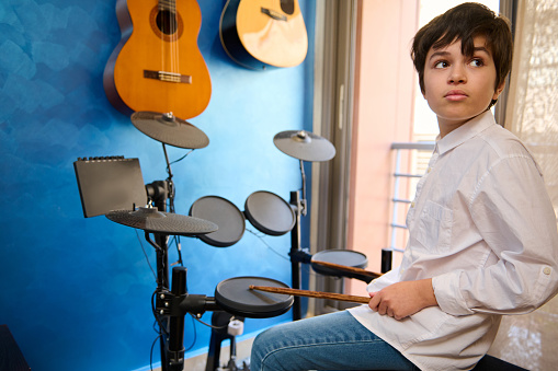 Confident Caucasian teen boy drummer musician sitting at drum set in a retro music studio at home, holding drumstick and dreamily looking aside. Electrical and acoustic guitars hanging on a blue wall
