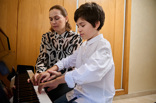 Handsome Hispanic boy in white casual shirt, learning playing piano, sitting at wooden piano forte and performing a musical composition under the guidance of is teacher. Musical education and hobby