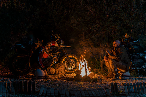 A man by a motorcycle at a campfire, resting his foot on the bike