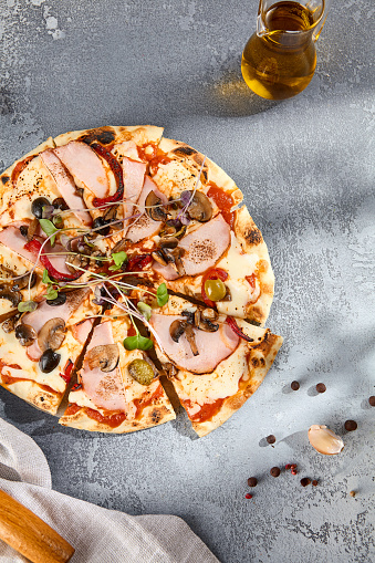 Top view of pizza with ham, mushrooms, and olives, artistically garnished with microgreens, on a gray textured surface.