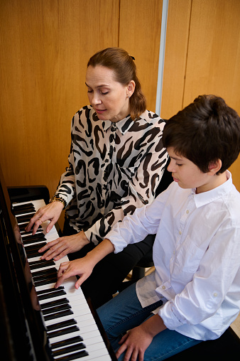 Vertical portrait of an authentic teenager boy having music lesson at home, sitting near his pianist teacher, playing grand piano, creating musical compositing and feeling the rhythm of sounds