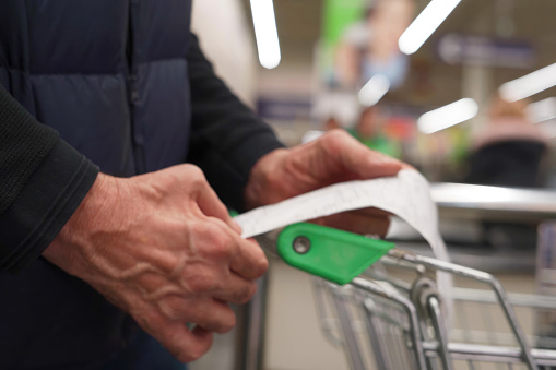 Buyer Checks Paper Receipt On Shopping Supermarket. The hands of an unrecognizable older man hold a sales receipt next to a grocery cart in a supermarket.