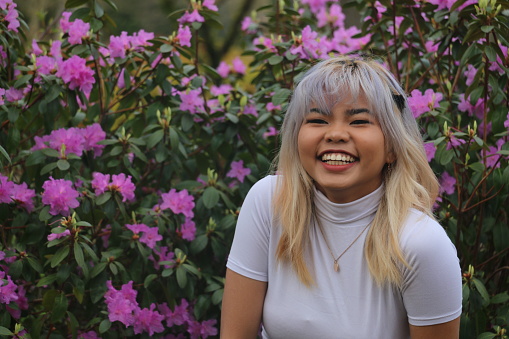 A closeup of a  Filipino model with a large smile while sitting in front of a pink rhododendron. She is wearing multi colored medium length hair, makeup, necklace and white tshirt.
