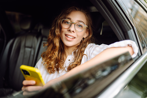 Young woman uses a smartphone while sitting in the back seat of a car. Female happy in car traveling on the road to destination. Technology, blogging, business concept.