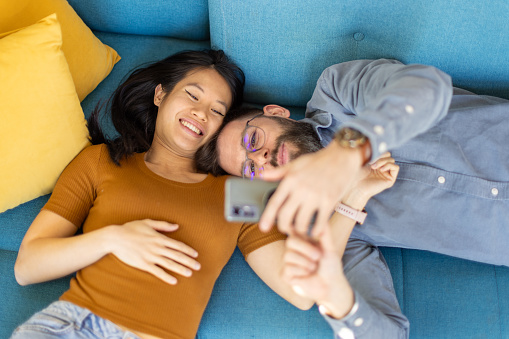 Mid adult interracial couple taking selfies while lying on sofa in their living room. They are smiling and look happy, carefree and relaxed.