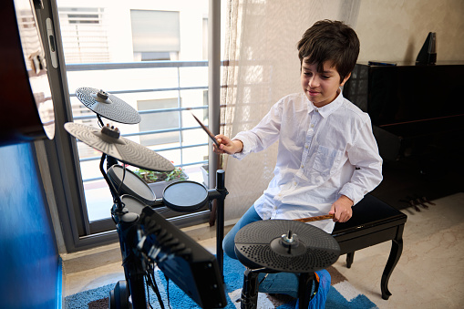 Handsome happy teenager boy playing drums at home music studio, holding drumsticks and creating music rhythm banging drums, beating on drum kit. Learning percussion musical instrument