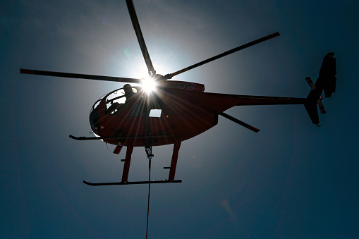 Fort Stockton, TX – November 6, 2020: An MD-500 helicopter is performing a variety of power line utility work and is silhouetted with a worker hanging from the side to perform the work.  The versatile MD-500 is the go to for power line work and it’s ability to be solid working base for utility workers.