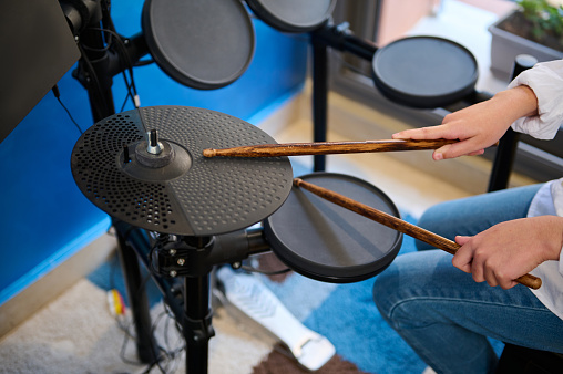 Close-up hands of teen boy musician creating rhythm of music while banging drums in stylish retro music home studio. Hands hold drumsticks and beating on black cymbals. Percussion musical instrument