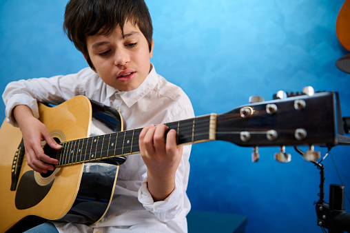Teenage boy pianist musician plucking the strings while playing the acoustic guitar. Authentic portrait of a confident adolescent boy learning to play guitar during a music lesson. Hobbies and leisure