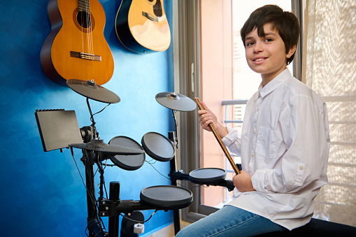 Teenager boy drummer in casual white shirt and blue jeans, sitting at drum set in his retro music studio , holding drumstick and smiling looking confidently at camera. Guitars hanging on a blue wall