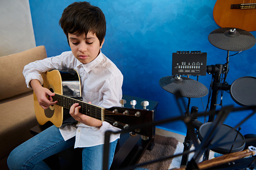 Confident portrait Hispanic teenage boy in white shirt and blue jeans, sitting in his blue studio room, playing acoustic guitar, focused on his music, filling the air with the sounds of his instrument