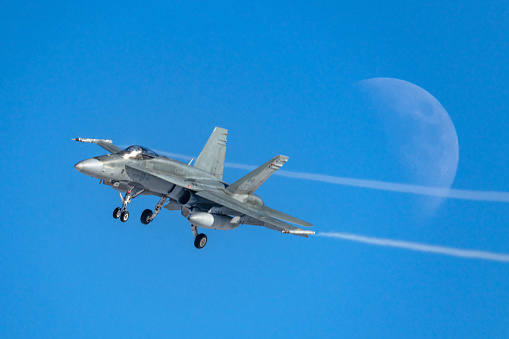 Bagotville, Quebec – January 17, 2024: A Royal Canadian Air Force FA-18 from RCAF Wing 3 is arriving at the Bagotville, Quebec, air base.  The landing FA-18 is creating some air flow lines as it banks for final approach to the runway in front of a rising moon.
