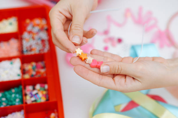 Woman making a bracelet of colorful beads with her hands. The woman hands holding beaded string and thread beads on it making the bracelet in studio. There are many colorful beads are on table with scissors.