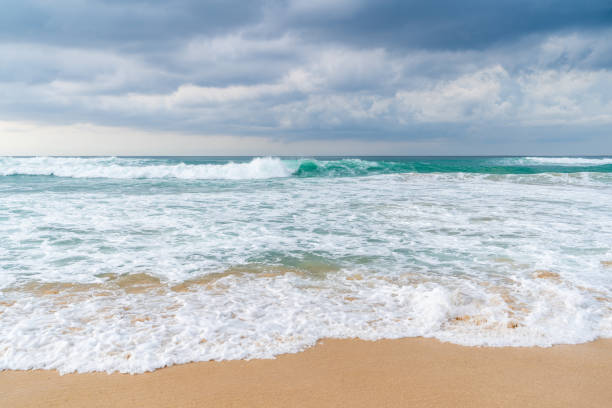 Bali, Dreamland - Waves crash onto the golden sandy beach, showcasing the vibrant turquoise color of the ocean under a dramatic cloudy sky, offering a visual spectacle of nature’s dynamic beauty and power.