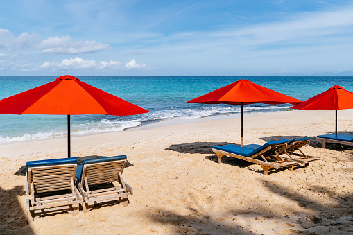 Red umbrellas and blue loungers are placed on the golden sands of a serene beach, offering a picturesque view against the backdrop of crystal clear waters and an expansive blue sky. Bali, Dreamland beach.