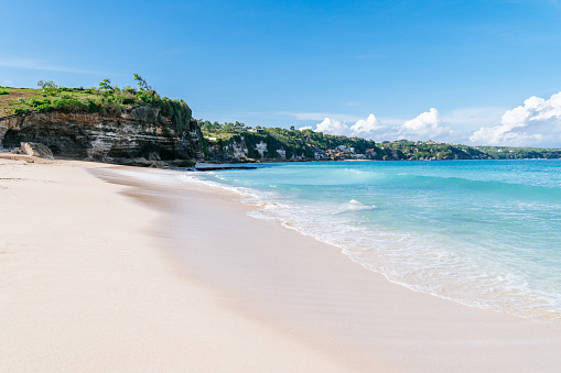 Bali - White sandy beach meets the clear blue waters of the ocean, with waves gently lapping at the shore; greenery-covered cliffs and distant hills provide a picturesque backdrop under a bright, cloud-speckled sky.