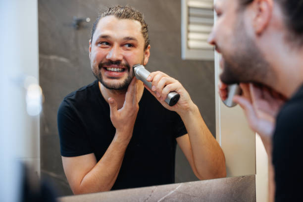 A man is using electric shaver to trim mustache and beard while standing in front of mirror in bathroom at home.