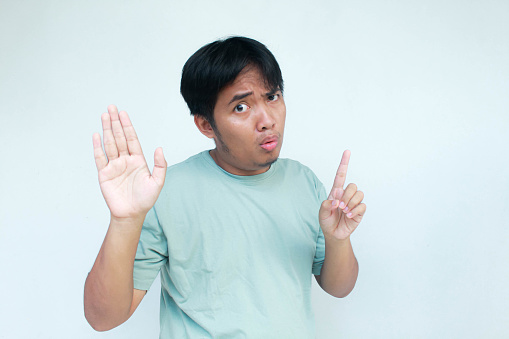 A young Asian man in a green t-shirt  looking unhappy and angry showing rejection and negative with thumbs down and stop or no gesture. Bad expression. Isolated in gray background.