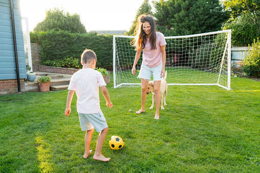 Young mother and son plays soccer with dog and have fun together. Happy family playing football with pet. Fun Playing Games in Backyard Lawn on Sunny Summer Day. Motherhood, childhood, togetherness
