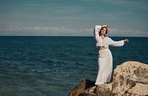 A beautiful woman in a white dress standing on a big rock in the middle of the sea.
