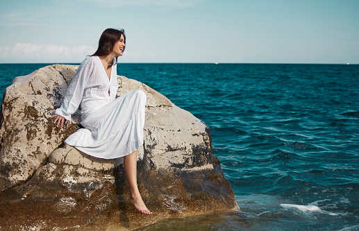 A beautiful woman in a white dress sitting on the rocks in the middle of the sea, playing with the water with her feet.
