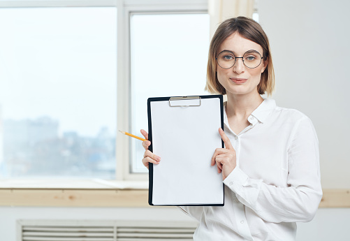 A woman is holding a folder with a white sheet of mockup paper and a window in the background. High quality photo