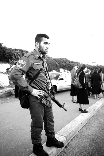 12-25-2014  Jerusalem .   Soldier  (on vacations) with machine gun  (TAR-21) of israeli Army in vacations  on bus stop or tram stop.  (also people on stop  - orthodox Judaist from behind). Monochrome photo