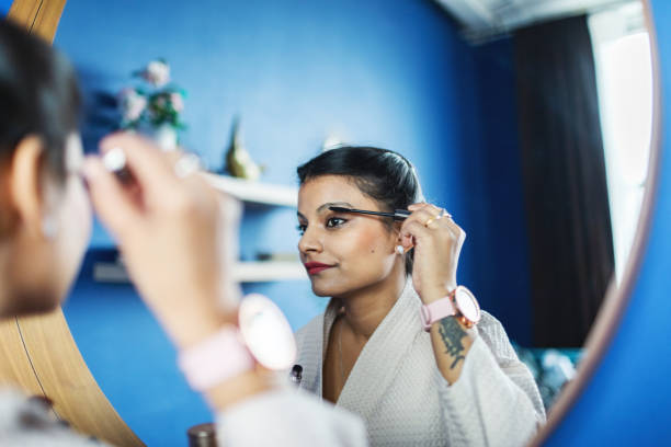 Young Indian woman in bathrobe looking at mirror and doing makeup