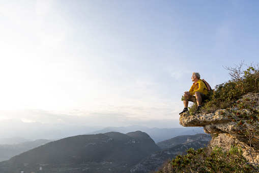 Mature hiker sits on cliff edge and looks out across distant mountain summits, Liguria