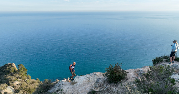 Mature couple ascend rock ridge crest above sea, Liguria