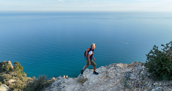 Mature man ascends rock ridge crest above sea, Liguria