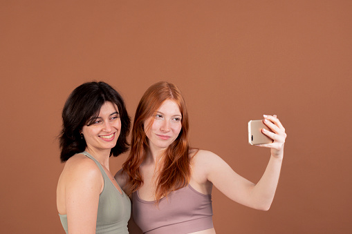 Two smiling women taking a selfie together against a brown background.