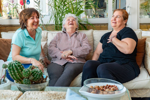 Happy elderly woman enjoying the affectionate bond of her daughter on Mother's Day. Two siblings, daughters of an elderly woman with grey hair, hugging, kissing, embracing female person and celebrating Mother's Day, Birthday, Eid Mubarak in living room. Domestic real live event. Family concept.