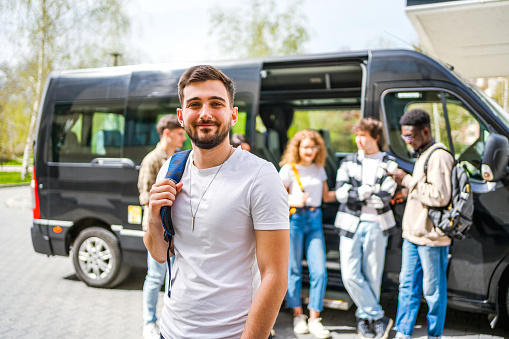 Portrait of a young cheerful male student in front of a minibus, ready for a trip. Traveling and tourism concept