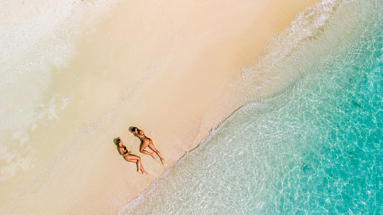 Aerial view of two gorgeous woman sunbathing and relaxing on the sand beach with beautiful crystal clear water. Perfect sunny day