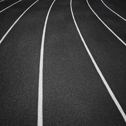 Overhead view of a rubber black running track surface with white lane lines. There is a slight curve of the white lane lines. Rubber black surface.
