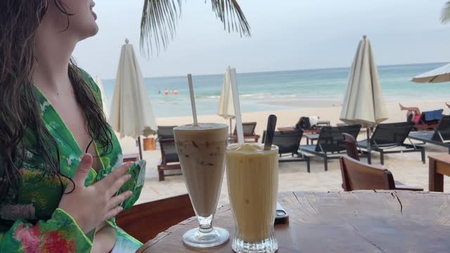 A girl with great delight drinks a coffee drink on the beach shows her fingers just perfect Okay she shows radiates the joy of life pleasure from relaxing in a green pareo with wet hair after swimming