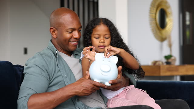 Black little girl saving coins in her piggy bank with the help of her loving father at home