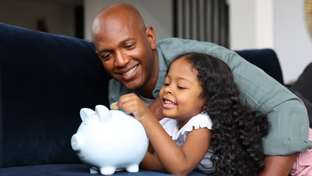 Loving African American father at home teaching his daughter to save money in a piggybank