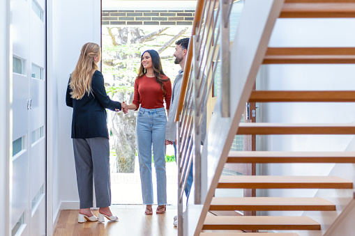 Real estate agent showing a young couple a new house. The home is contemporary. All are happy and smiling. The couple are casually dressed and the agent is in a suit and shaking hands .The couple are being greeted at the front door by the real estate agent.