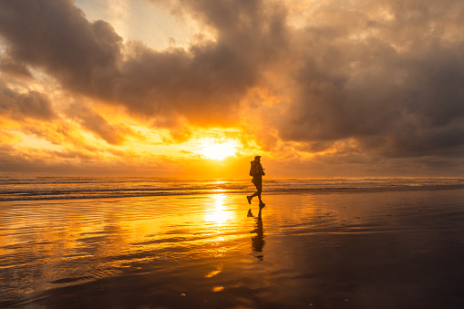 Active senior Asian man enjoying a run out at the sea in casual clothing with reflection in water at sunset.