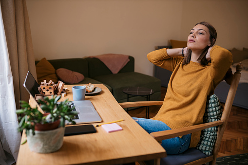 Pretty young woman taking break while working from home. Beautiful female freelance sitting at desk looking away and thinking with her hands behind head.