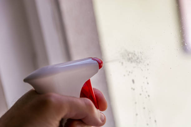 Captured in this close-up shot, a hand expertly grips a window cleaning spray bottle against the backdrop of a window. The focus is on the action of the index finger pressing the nozzle, releasing a stream of cleaning solution onto the glass surface. This dynamic image showcases the motion of the spray, with droplets mid-air, highlighting the ease and effectiveness of using spray cleaners for spotless windows. The transparent window allows natural light to filter through, illuminating the fine mist, and emphasizing the cleaning process in action. This visual not only demonstrates the practical use of cleaning tools but also celebrates the simple yet impactful act of maintaining cleanliness and clarity in our surroundings.